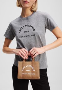Grå bomulds-t-shirt med sort tekst, kombineret med en lille brun lærreds-taske med hvidt logo. Enkel, afslappet stil.