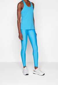 Osoba w jasnoniebieskich legginsach sportowych i pasującym topie na ramiączkach, w połączeniu z białymi i szarymi butami biegowymi, stoi na białej podłodze.