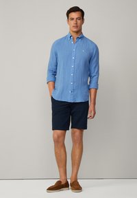 Chemise en lin bleu clair avec col boutonné, associée à un short marine et des mocassins en daim marron. Coupe simple et décontractée.