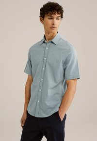 Šviesiai mėlynas trumpų rankovių marškinėliai su švelniu tekstūriniu raštu, apykakle ir baltais mygtukais. Dėvimi su tamsiomis kelnėmis.