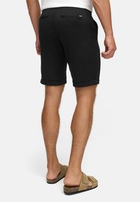 Schwarze Shorts mit umgeschlagenen Säumen, aus weichem Material gefertigt. Verfügt über zwei Gesäßtaschen und ein Logodetail. Kombiniert mit beigen Slipper-Sandalen.