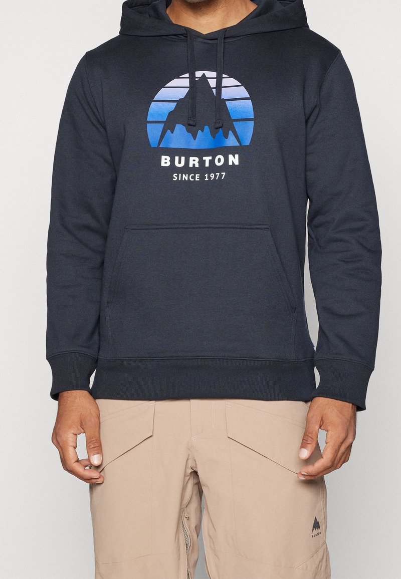 Homme portant un sweat à capuche Burton noir avec un motif de montagne bleu et un pantalon beige avec des poches avant visibles.