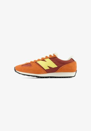 Niedriger Sneaker aus orangefarbenem Wildleder, rötlich-braunem Stoff, weißen Schnürsenkeln und einem großen gelben Seitenlogo auf einer weißen Sohle.