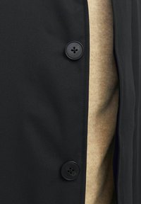 Černá vnější bunda s hladkou texturou, s kulatým černým knoflíkem a béžovým vnitřním oděvem, který je částečně viditelný.