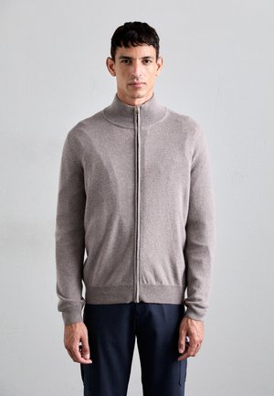 Lysebrun zip-up trøje lavet af blødt strikmateriale. Har en høj krave og ribbede manchetter og kant. Båret med mørke bukser.