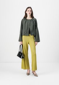 Veste cropped vert olive avec encolure froncée, associée à un pantalon large côtelé moutarde. Le mannequin tient un petit sac à main noir et porte des talons gris.