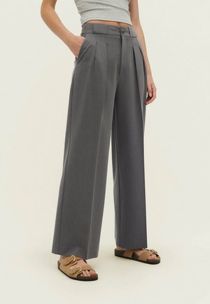 Persona con pantalones grises de talle alto y pierna ancha con pliegues, una camiseta corta gris y sandalias marrones con hebillas.