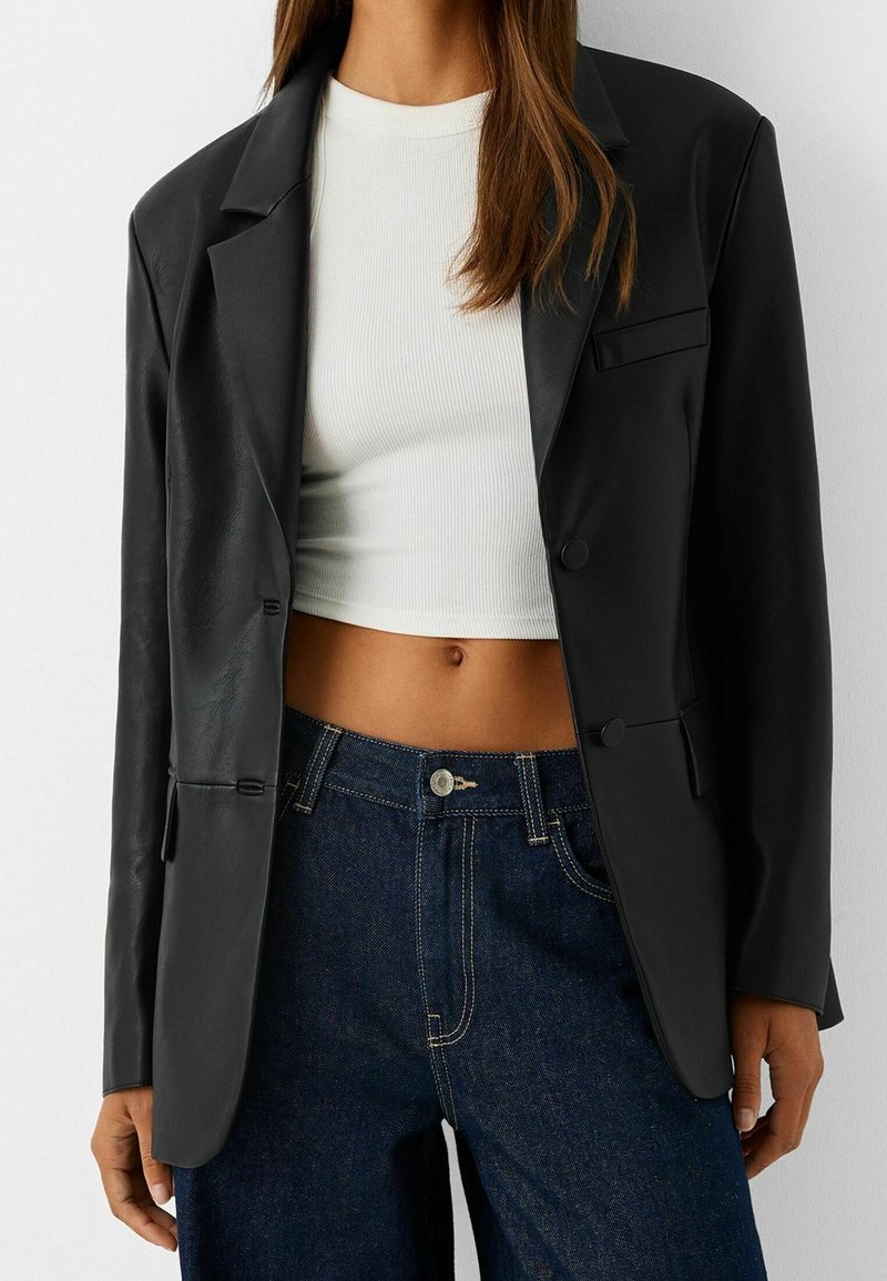 Femme portant un blazer en cuir noir sur un crop top blanc côtelé et un jean bleu foncé taille haute.