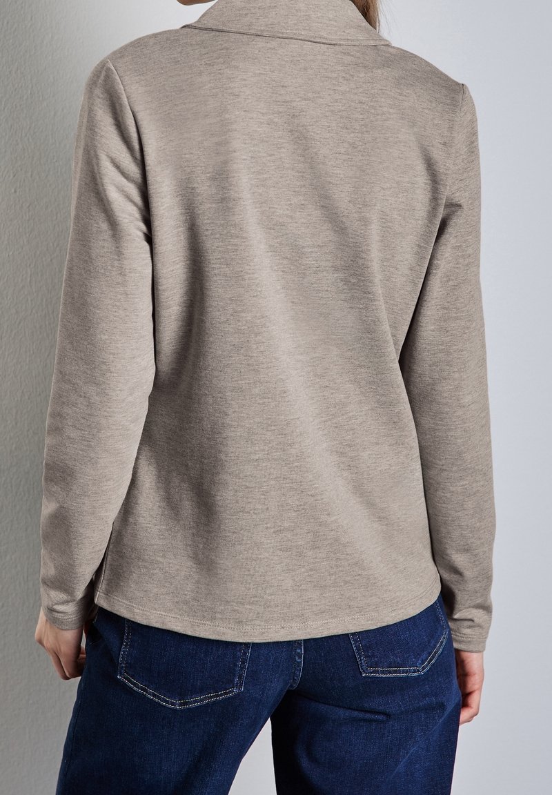 Personne portant un haut à manches longues beige clair et un jean en denim bleu foncé, vue de dos devant un mur uni.