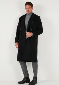 Cappotto doppiopetto in lana nera, indossato sopra un dolcevita grigio. Pantaloni grigi aderenti e stivaletti neri completano il look.