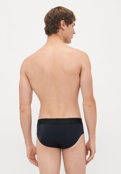 Junger Mann, von hinten gesehen, der schwarze Badehose mit einem breiten Bund trägt, vor einem schlichten weißen Hintergrund stehend.