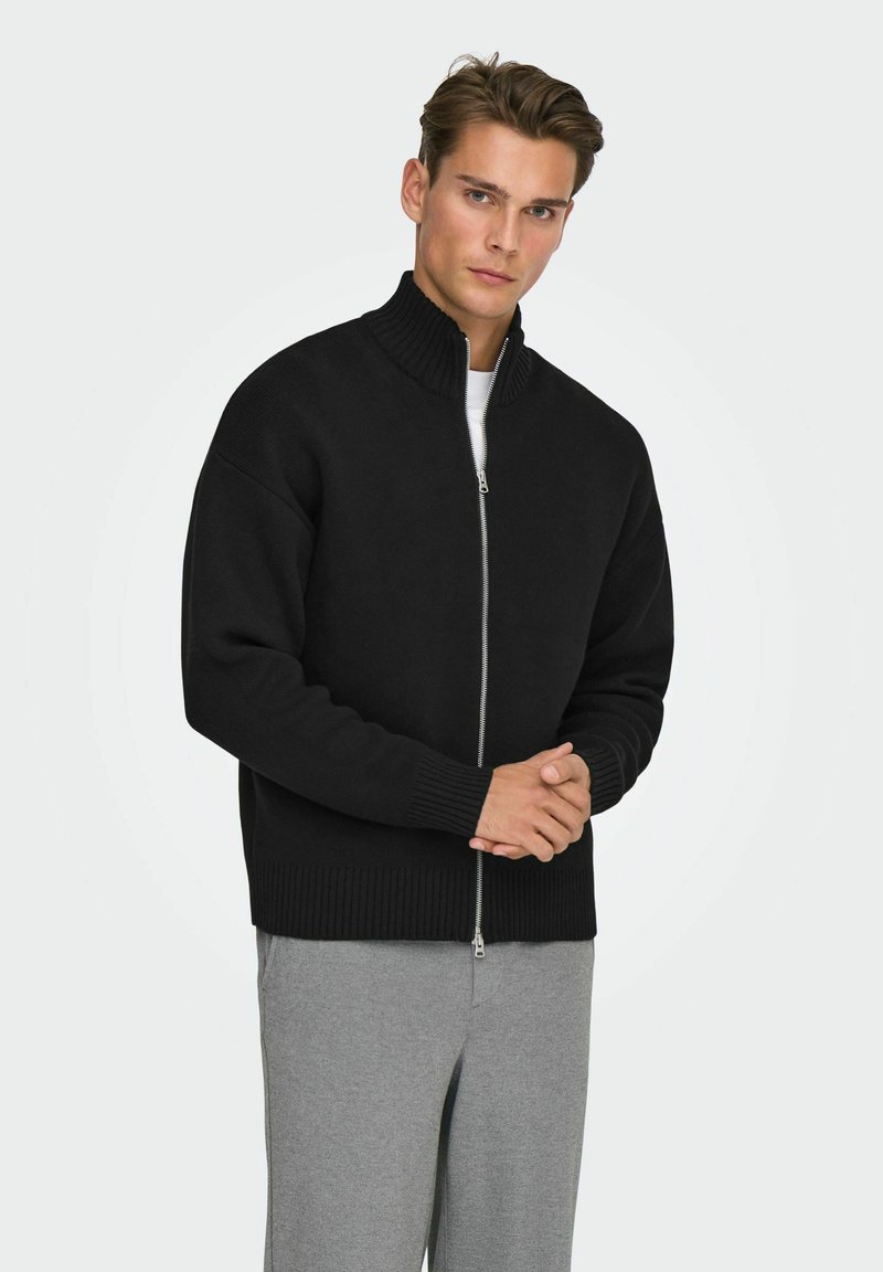 Pull noir à fermeture éclair avec col montant, poignets et ourlet côtelés. Porté par-dessus une chemise blanche, associé à un pantalon gris clair. Texture lisse.