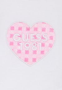 Λευκό ύφασμα με ροζ, λαμπερή καρδιά με μοτίβο καρό. Κείμενο «GUESS 1981» σε λευκά ανάγλυφα γράμματα μέσα στην καρδιά.