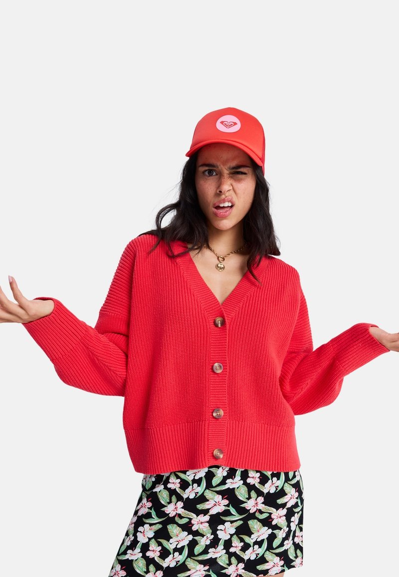 Mladá žena v červenej šiltovke a svetri, s kvetovanou sukňou, krčí ramenami a má zmätený výraz na tvári, na jednoduchom pozadí.