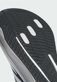 Suola di scarpa in gomma nera con motivo di battistrada in rilievo, dettagli bianchi, presenta tre intagli ovali e un'area grigia strutturata vicino al bordo.