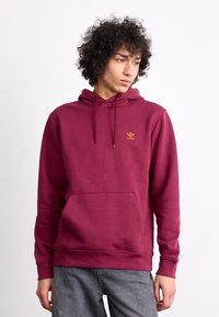Burzgino spalvos džemperis, pagamintas iš švelnios medžiagos, su kišenių kanaguru, suvirkimo kapuce ir mažuoju auksiniu adidas logotipu ant krūtinės.