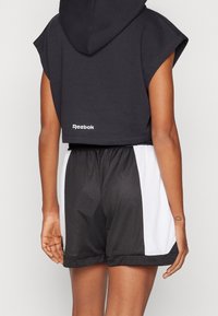 Felpa nera corta con cappuccio, con logo Reebok bianco. Abbinata a pantaloni corti sportivi in rete neri e bianchi.