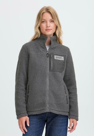 Femme portant un pull en polaire gris zippé avec une poche poitrine et un jean bleu, debout devant un fond clair uni.