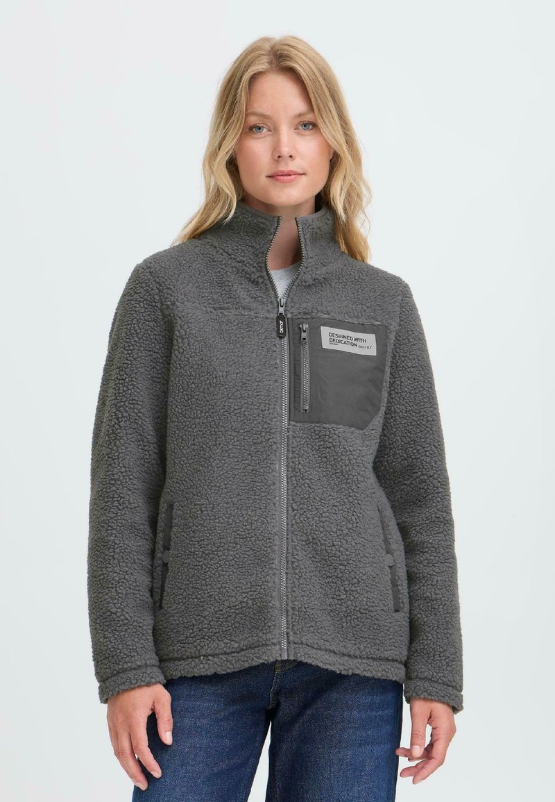 Kvinne iført grå zip-up fleecejakke med brystlomme og blå jeans, står mot en enkel lys bakgrunn.