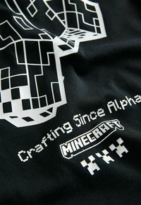 Tessuto nero con design di teschio a pixel bianchi, "Crafting Since Alpha", logo di Minecraft e tre piccoli personaggi a pixel sotto il testo.