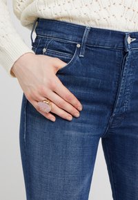 Zlatý a růžový prsten se kamenným akcentem nasazený na ruce, vedle něj blízký záběr na tmavě modré džíny s jemnými detaily opotřebení.