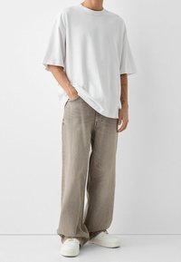 Camiseta blanca de gran tamaño con mangas cortas, combinada con pantalones de mezclilla de pierna ancha en un tono beige claro y zapatillas blancas. Conjunto casual con un ajuste relajado.