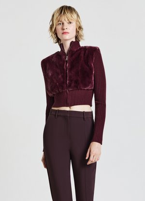 Maglione corto bordeaux con parte anteriore in pelliccia sintetica e maniche a coste, abbinato a pantaloni sartoriali bordeaux scuro. Chiusura con zipper al collo.