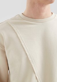 Beige katoenen sweatshirt met een rafelrand detail dat diagonaal over de borst loopt, met een geribde halslijn en een gladde textuur.
