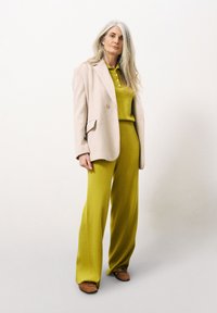 Blazer beige ajusté sur un haut en tricot vert vif et un pantalon large vert. Des ballerines marron complètent la tenue.