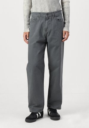 Personne portant un pantalon ample gris foncé, un haut à manches longues côtelé gris clair et des baskets noires avec des rayures blanches, debout sur un sol blanc.