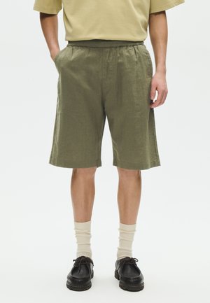 Person iført olivengrønne shorts, beige skjorte, cremefarvede sokker og sorte sko, der står med hænderne i lommerne på en hvid baggrund.