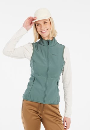 Grüne Softshell-Weste mit hohem Kragen und Reißverschlüssen, über einem cremefarbenen Langarmoberteil getragen. Eine hellfarbene Kappe und braune Hosen runden das Outfit ab.