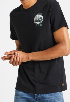 Mann trägt schwarzes G-Star Raw T-Shirt mit Logotext auf der Brust und blaue Jeans, die Hände vor dem Körper verschränkt.