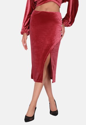 Kvinde iført en rød velour slå-om-nederdel med sidelomme og dekorative knapper, kombineret med en matchende top og sorte højhælede sko.