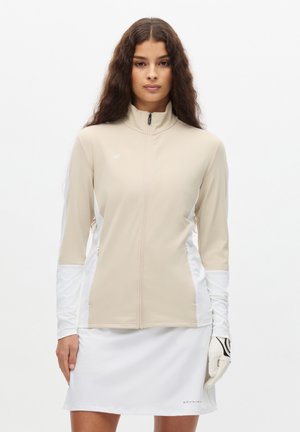Femme aux longs cheveux bouclés portant une veste beige zippée, un gant de golf blanc et une jupe blanche sur fond blanc.