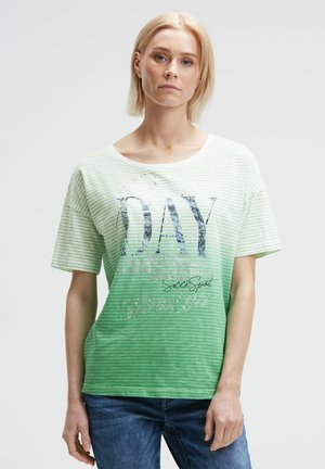 Blonde Frau trägt ein grün-weiß gestreiftes T-Shirt mit dem Aufdruck "DAY", kombiniert mit blauen Jeans, die vor einem hellen Hintergrund steht.