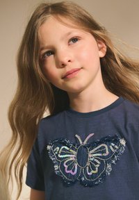 Mörkblå t-shirt med en stor fjäril designad av paljetter och slitna tygdetaljer. Korta ärmar och rund halsringning.