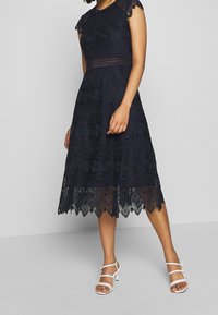 Marinblåspetsklänning med figurnära överdel, korta ärmar och en utsvängd kjol med en scallopad nederkant. Kombinerad med vita remsandaler.