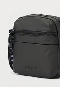 Μικρή μαύρη ορθογώνια τσάντα ώμου με μπροστινή τσέπη με φερμουάρ, με το λογότυπο "HUGO" εγχάρακτο στο κάτω μπροστινό πάνελ και ρυθμιζόμενο λουρί με σχέδιο.