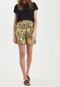 Musta pistekuvioinen t-paita, keltaiset kukkakuosiset shortsit mustilla yksityiskohdilla. Malli käyttää ruskeita litteitä sandaaleja. Yksinkertainen valkoinen tausta.