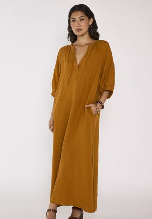 Femme aux longs cheveux foncés portant une robe maxi ample couleur moutarde avec manches trois-quarts, poche latérale et talons bruns, debout.