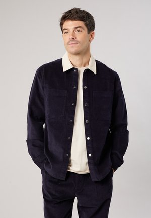 Man draagt een donkerblauwe corduroy jas met een witte kraag en bijpassende broek, staat met handen in de zakken en kijkt iets naar de zijkant.