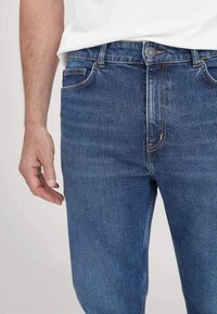 Blauwe denim jeans met een rechte snit, vijf zakken en stikte accenten. De stof heeft een gladde textuur en een lichte vervaging.