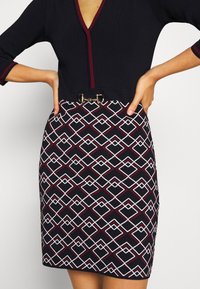 Femme portant une robe noire à col en V profond, manches 3/4, et une jupe à motif géométrique avec une boucle dorée décorative à la taille.