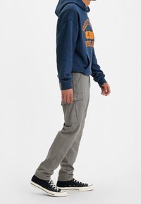 Grå cargobyxor med flera fickor, tillsammans med en marinblå hoodie med orange text. Svarta sneakers med vita sulor avslutar looken.