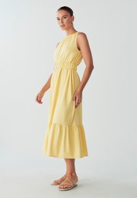 Vestito giallo senza maniche con vita arricciata, orlo a campana plissettato e tessuto di cotone morbido. Indossato con sandali piatti color talpa.