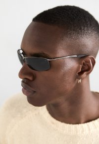 Lunettes de soleil avec des verres foncés, des montures rectangulaires fines et des accents argentés. Portées par une personne dans un pull léger, vue de profil en gros plan.