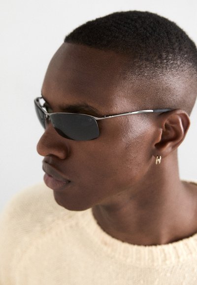 Lunettes de soleil avec des verres foncés, des montures rectangulaires fines et des accents argentés. Portées par une personne dans un pull léger, vue de profil en gros plan.