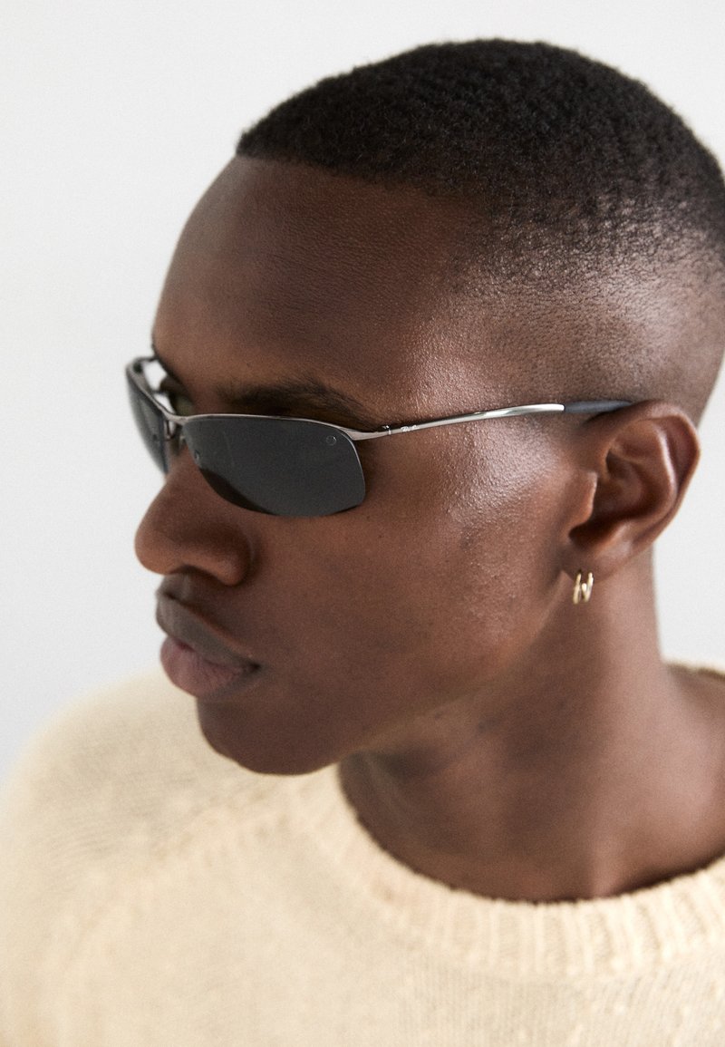 Lunettes de soleil avec des verres foncés, des montures rectangulaires fines et des accents argentés. Portées par une personne dans un pull léger, vue de profil en gros plan.