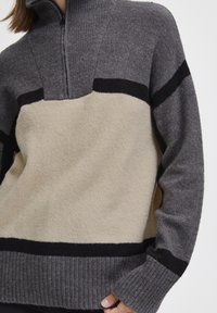 Pull gris et beige avec un col haut, doté d'une fermeture à demi-zip. Mélange de laine texturée avec des rayures horizontales noires et des poignets côtelés.